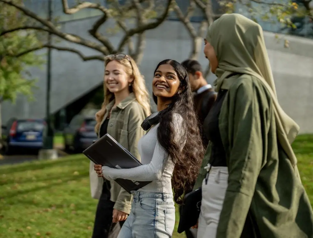 A group of university students are seen walking outside on campus as they make their way from residence to class. They are each dressed casually and have backpacks and book bags with them.