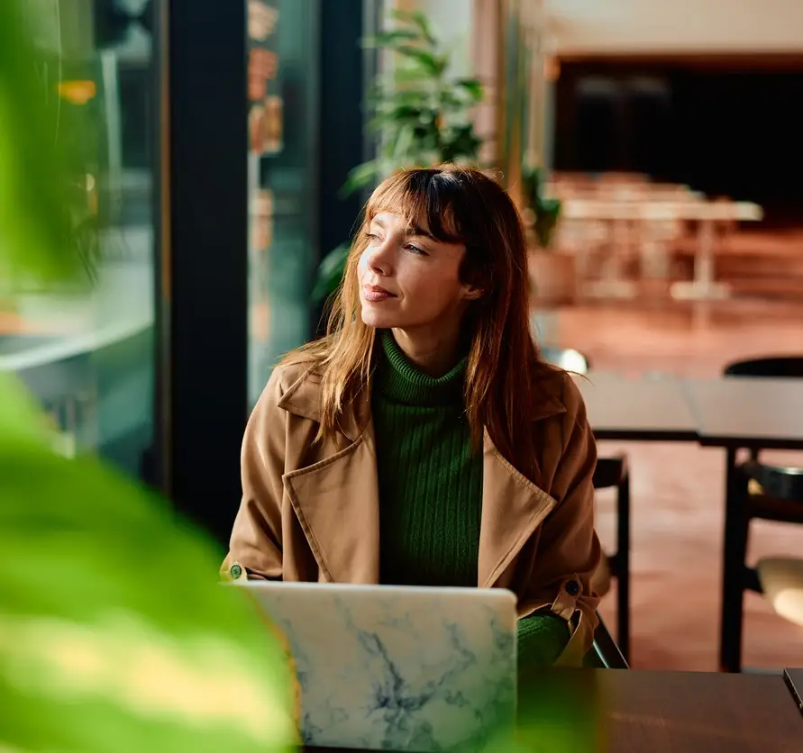 Woman freelancer taking a break from work, sitting at a cafe table with her laptop, contemplating while looking away with sunlight streaming through the window